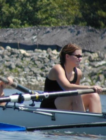 A white woman with short brown hair and sunglasses in a black singlet holding an oar in a crew shell.