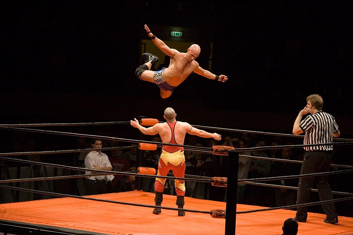 A wrestler in just shorts and kneepads is suspended in midair, while another man in a red and yellow singlet prepares for him to fall on top of him. A referee waits in another corner. 