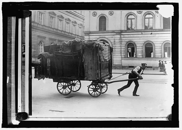 A black and white photo of a man in a vest and hat pulling a large wagon full of clothes and furniture., set in the early 1900s.