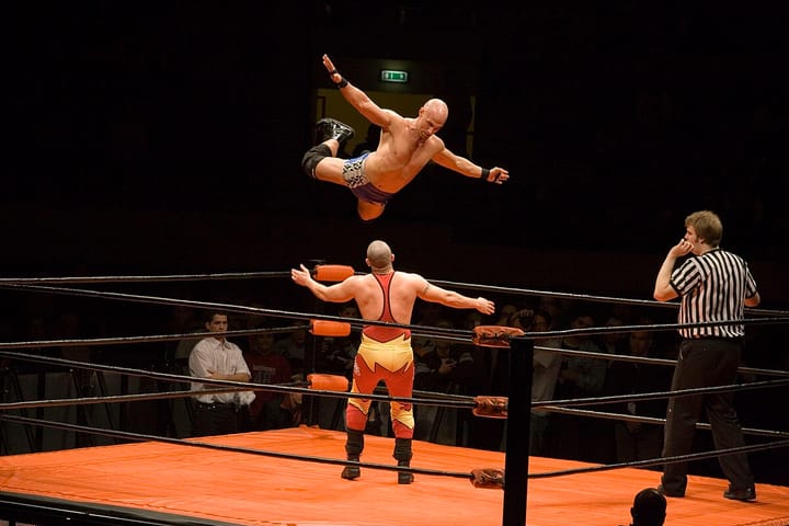A wrestler in just shorts and kneepads is suspended in midair, while another man in a red and yellow singlet prepares for him to fall on top of him. A referee waits in another corner.