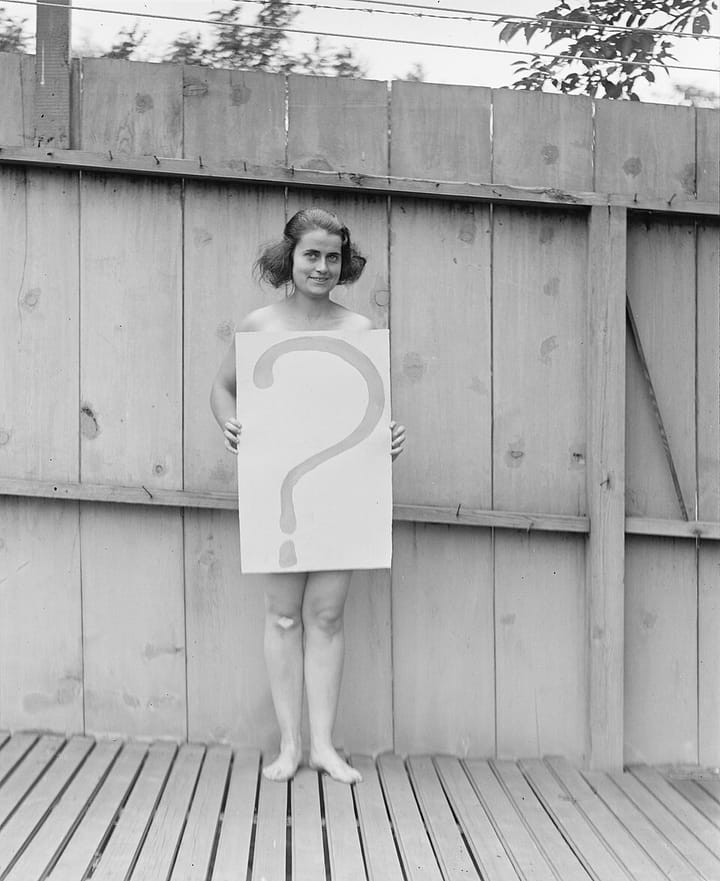 A black and white photo of a white woman with dark hair holding a sign with a question mark on it covering her torso.