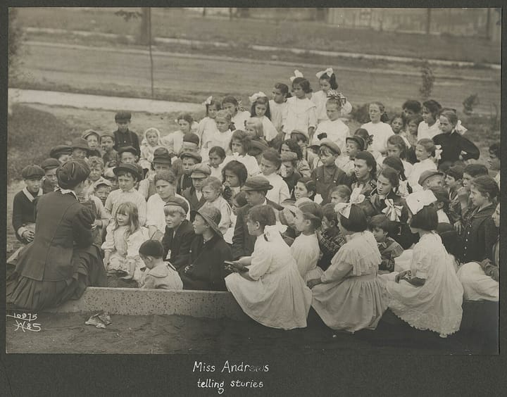 A black and white image from 1910 of a female teacher with her back to the camera reading a book to a large number of white children. The photo is captioned "Miss Andrews telling stories."