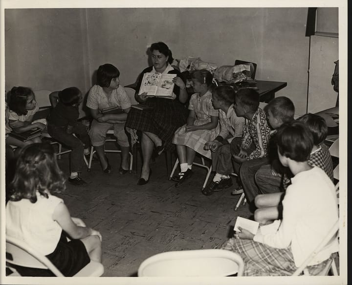 A black and white picture of students sitting in a chairs in a circle while their teacher reads to them.
