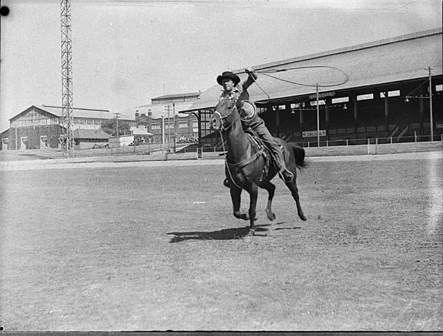 A black and white photo of a man in a cowboy hat riding a course and throwing a lasso.