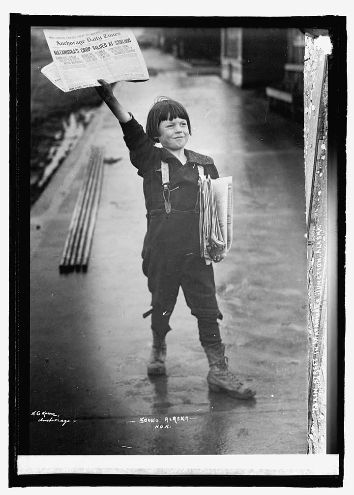 A young white boy in short pants and suspenders holding a newspaper in the air with one hand and a stack to sell in the other. 