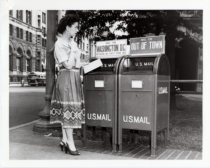 A black and white photo of a white woman in a long skirt, holding a letter, deciding which mailbox to put it in. The mailboxes are labeled Washington, DC and Out of Town, and U.S. Mail.