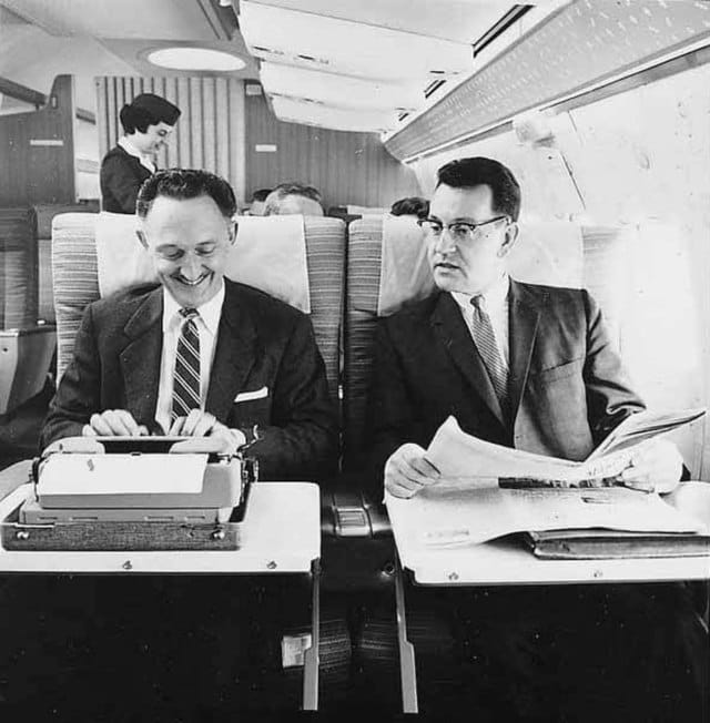 Two men on Northwest Airlines aircraft, one using typewriter, with female flight attendant in background, ca. 1965